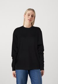 Črn pulover s crewneck izrezom in dolgimi rokavi, izdelan iz mehkega blaga. Oblikovanje vključuje sproščen kroj ter rebraste manšete in rob, v kombinaciji z modrimi kavbojkami.