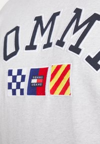 Maglietta grigia in cotone con grandi scritte ricamate in blu navy e tre toppe colorate: a scacchi, a righe rosse e design giallo.