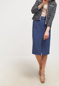 Mörkblå pennkjol med fram slit och två fickor, kombinerad med en strukturerad grå jacka, bärs med ljuslila sandaletter med remmar.