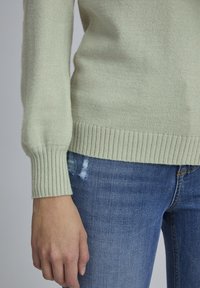 Lys grøn strikket sweater med ribbede manchetter og kant, parret med slidte blå jeans. Blød tekstur, tætsiddende design.