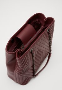Sac en cuir matelassé bordeaux avec accents en chaîne, fermeture éclair et pochette intérieure amovible. Présente une texture lisse et une forme structurée.