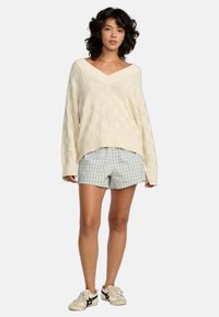 Cremfarbener Strickpullover mit V-Ausschnitt und kariertem Muster, kombiniert mit hellblauen Karoshorts. Weiße Sneakers mit schwarzen Akzenten.