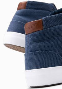 Zapatillas de lona azules con un tirante de cuero marrón en el talón, suela de goma blanca, tela texturizada y un diseño minimalista.