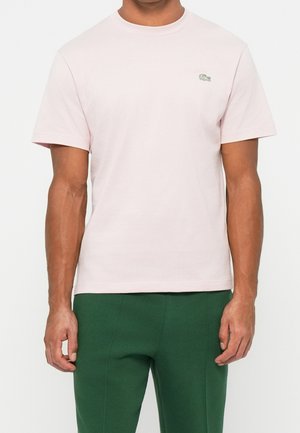Man draagt een lichtroze T-shirt met korte mouwen met een klein groen logo op de borst en donkergroene broek, staand met ontspannen armen.