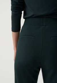 Ciemnozielone spodnie o gładkiej fakturze materiału, z dwiema tylnymi kieszeniami i dopasowanym paskiem w talii. Design jest prosty i skrojony.