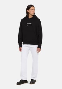 Sudadera negra con capucha, con un bolsillo frontal y texto de logo en blanco. Combinada con pantalones blancos y zapatos negros, con punta redonda y tacón bajo.