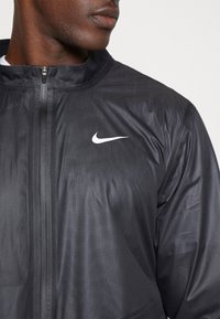 Sort letvægtsjakke med lynlås krave, der har et reflekterende hvidt Nike-logo på venstre bryst og glat, tekstureret stof.
