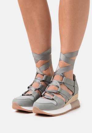 Baskets de sport grises avec des courroies croisées, matériau texturé, semelle en coin et semelle en caoutchouc. Présentent un accent tissé au niveau du talon.