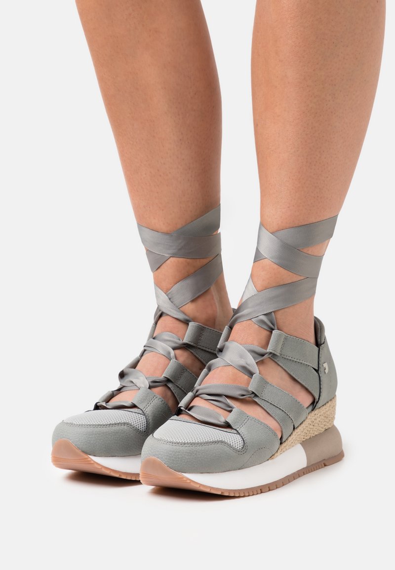 Zapatillas deportivas grises con correas cruzadas, material texturizado, suela en cuña y suela de goma. Presentan un detalle tejido en el talón.