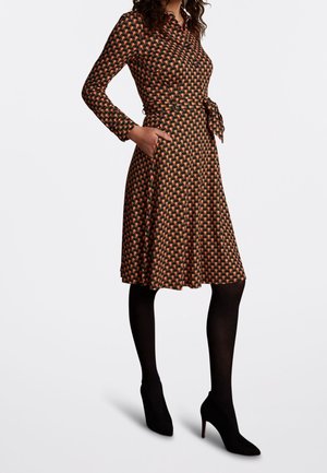 Woman wearing a long-sleeve, patterned wrap dress with a tie waist, paired with black tights and black high-heeled shoes.