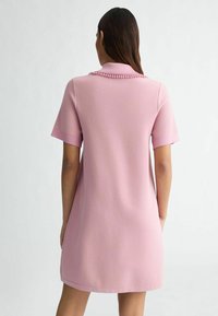 Robe rose à manches courtes avec un col, présentant un tissu texturé. Le col possède des accents décoratifs et la robe a une coupe décontractée.