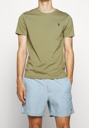 Homme portant un t-shirt à manches courtes vert olive avec un logo bleu marine et un short décontracté bleu clair, debout avec une main dans la poche.