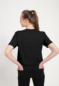 Zwart T-shirt met korte mouwen en een ronde hals, gemaakt van katoen. Achteraan getoond, gecombineerd met een donkere spijkerbroek, handen in de zakken.