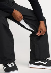 Pantalons cargo noirs avec des évents latéraux zippés, dotés d'une doublure en maille. Associés à des baskets noir et blanc, montrant un tissu texturé.