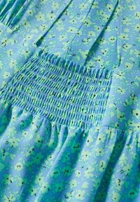 Gros plan d'un tissu bleu clair avec de petites fleurs vertes et blanches et un détail de couture smockée horizontale.
