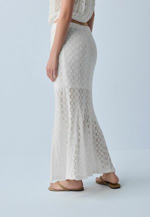 Frau trägt einen weißen Spitzen-Maxirock und passenden Crop-Top, steht barfuß mit gebräunten Flip-Flops vor einem hellblauen Hintergrund.