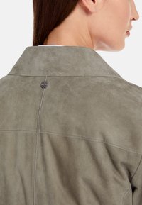 Gray suede jacket with a smooth texture, featuring a wide collar, visible stitching, and a branded metal snap at the back.