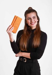 Oranžová peněženka s texturovaným povrchem, s elegantním designem a detaily loga. Drží ji osoba oblečená v černém dlouhém rukávu.