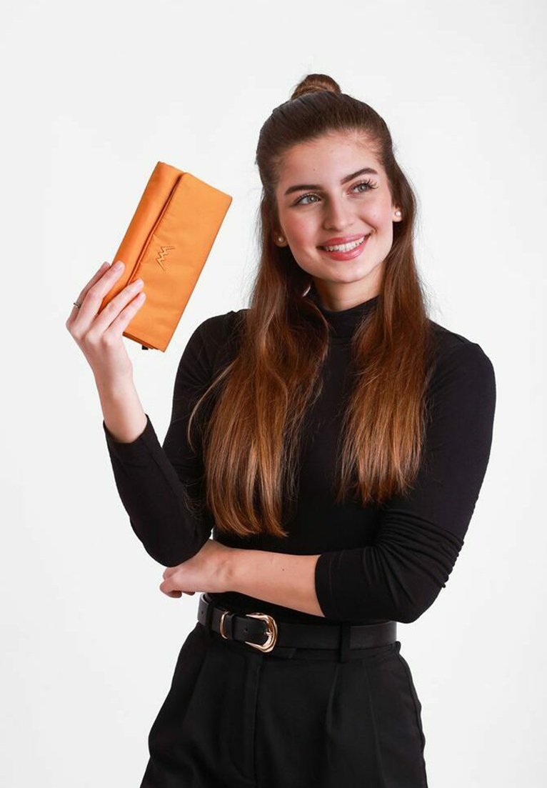 Oranžová peněženka s texturovaným povrchem, s elegantním designem a detaily loga. Drží ji osoba oblečená v černém dlouhém rukávu.