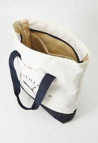 Witte stoffen tote bag met marineblauwe accenten en logo. Twee stevige handvatten en een gevoerde binnenkant met vakken. Ontwerp met open bovenkant.