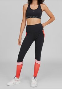 Czarny stanik sportowy z logo na pasku. Legginsy z wysokim stanem w kolorze czarnym z czerwonymi i różowymi akcentami. Sneakersy z białą podstawą i kolorowymi detalami.