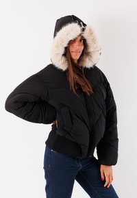 Giubbotto puffer nero con cappuccio in pelliccia sintetica rimovibile, polsini a coste e due tasche frontali. Il motivo trapuntato aggiunge una texture e un dettaglio di design.
