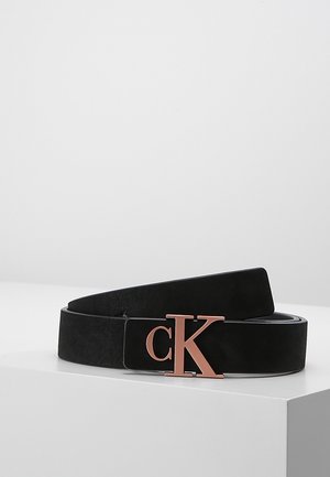 Černý kožený pásek s přezkou se symbolem "cK" v růžovém zlatě, stočený na bílém povrchu na jednobarevném světle šedém pozadí.