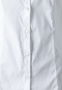 Gros plan sur le devant d'une chemise blanche montrant la texture du tissu et cinq boutons blancs fixés en rangée verticale.