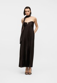 Femme debout portant une robe maxi noire plissée sans bretelles avec une petite découpe à l'avant, un foulard noir autour du cou et des talons ouverts noirs.