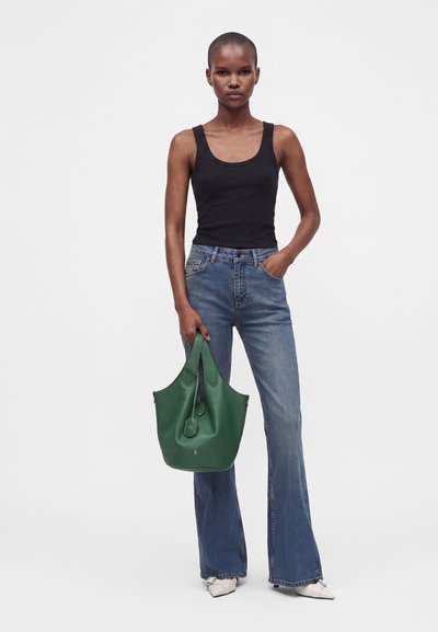 Femme portant un débardeur noir et un jean bleu tient un grand sac à main vert d'une main, l'autre main dans la poche, debout sur des chaussures blanches.