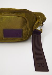 Olive green bag with a crinkled texture, featuring a zip pocket and a purple logo patch. Strap has vertical grooves and a clip attachment.