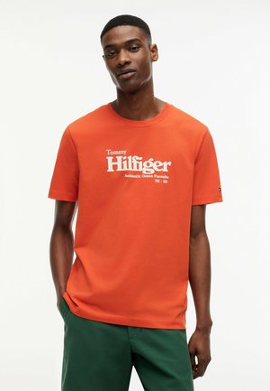 Joven vestido con una camiseta naranja de Tommy Hilfiger con texto de logo blanco y pantalones verdes, de pie frente a un fondo blanco.