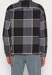 Harmaa-musta ruudullinen flanellipaita, jossa napitettava edessä, pitkät hihat ja sivuhalkiot. Paita esittelee erilaisia sävyjä ja tekstuureja.