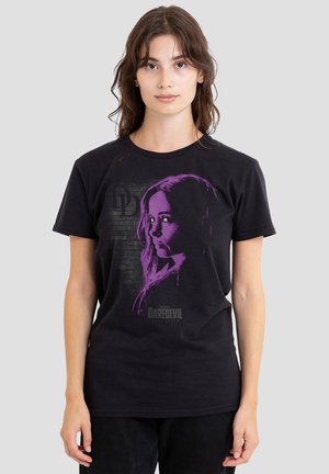 Young woman standing facing forward wearing a black Daredevil t-shirt with purple graphic portrait of a female character.