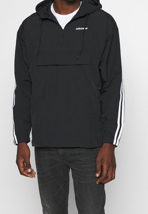 Sort Adidas hættetrøje med hvide striber på ærmerne og halvlukning med lynlås, båret over en hvid skjorte med sorte jeans.