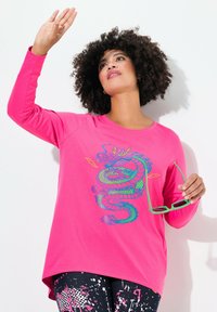 Rosa Langarmshirt mit einem bunten Drachenmotiv; kombiniert mit gemusterten Leggings. Model hält grüne Sonnenbrille und hebt die Hand.