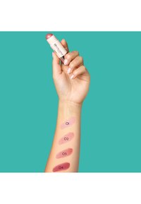 Stick de blush avec un dessus arrondi tenu dans une main ; échantillons de quatre teintes en dégradé sur un avant-bras, étiquetés de O1 à O4. Fond aqua lumineux.