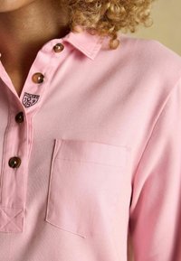 Heleda roosa pluus, millel on kraega kaelus, pruuni nööpide, rinnatasku ja selle kohal väike logo. Sujuv, tekstureeritud kangas.