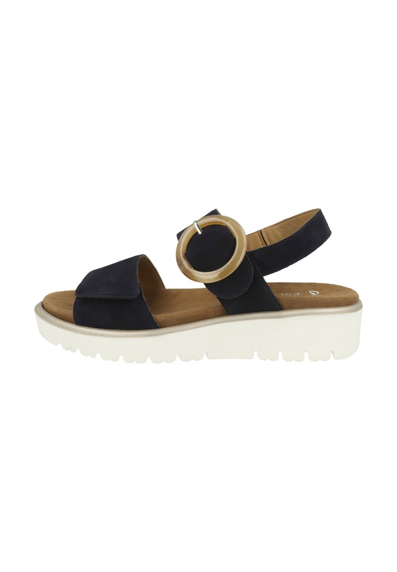Sandalo in suede blu navy con una larga fascetta anteriore, una fascia posteriore regolabile e un grande accento circolare. Presenta una suola bianca e texturizzata.