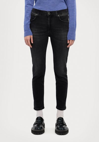 Jeans en denim noir à coupe slim, avec cinq poches, fermeture à bouton et détails de surpiqûre subtils. Portés avec des mocassins noirs brillants et des chaussettes blanches.