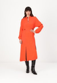 Heldere oranje ritsjurk met lange mouwen, voorzien van een split aan de voorkant en een trekkoorddetail, gedragen met zwarte laarzen tot de knie. Glad textuur.