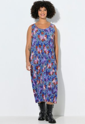 Smilende kvinde med krøllet hår iført en ærmeløs blå blomsterkjole og sorte støvler til midt på læggen, stående mod en hvid baggrund.