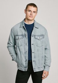 Lichtblauwe denimjack met twee borstzakken, knopen aan de voorkant en subtiele stikdetails. Draag het over een donkergroene geribde trui.