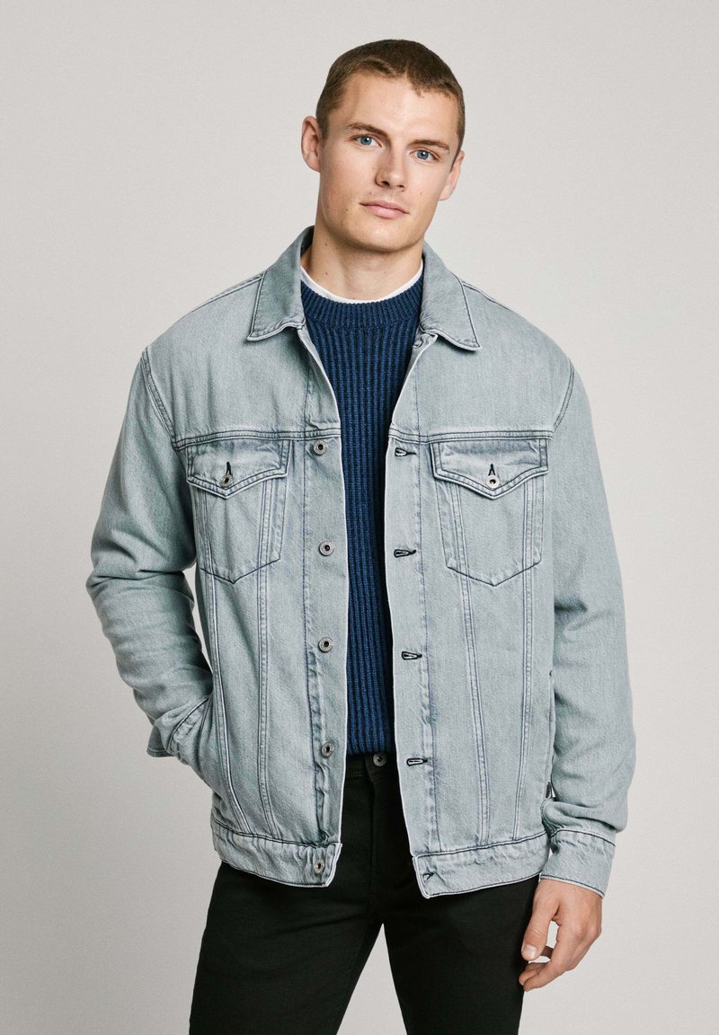 Lichtblauwe denimjack met twee borstzakken, knopen aan de voorkant en subtiele stikdetails. Draag het over een donkergroene geribde trui.