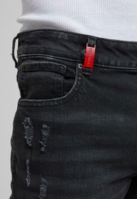 Jean noir usé porté avec un haut côtelé blanc, avec une étiquette rouge attachée à une passoire de ceinture du côté droit.