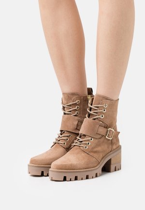 Bottines en suède marron avec une semelle épaisse, devant lacé et détail de sangle à boucle. Comprend des œillets métalliques et une finition texturée.