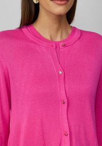 Cardigan rose vif avec un col rond, texture nervurée et petits accents de boutons brillants sur le devant.