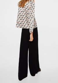 Blouse met een geometrisch patroon in roze, zwart en beige, gecombineerd met zwarte wijde broekspijpen. De blouse heeft lange mouwen en een losse pasvorm.