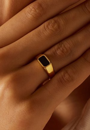 Bague en or avec une pierre noire rectangulaire incrustée. Surface lisse et polie. Portée sur une main avec une texture de peau visible.