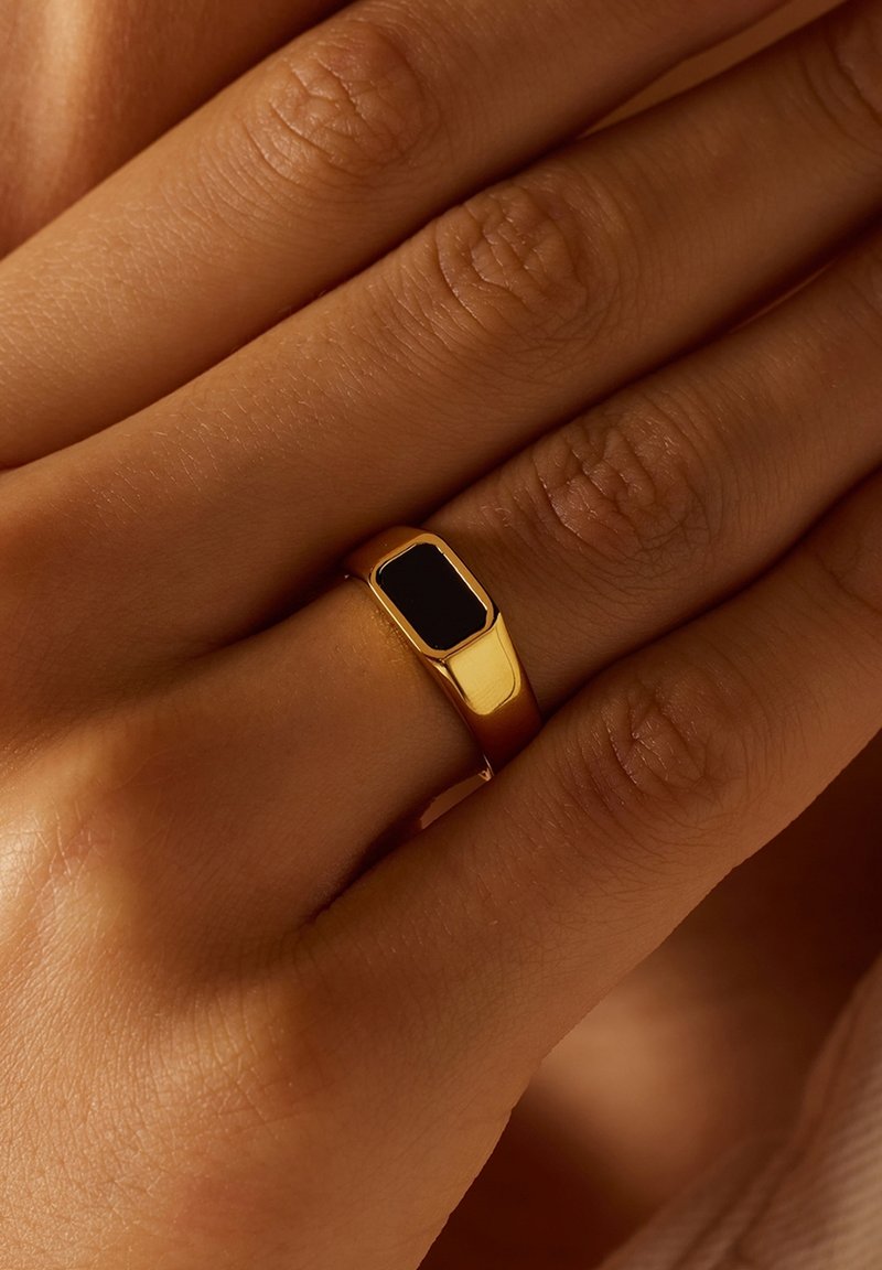 Bague en or avec une pierre noire rectangulaire incrustée. Surface lisse et polie. Portée sur une main avec une texture de peau visible.